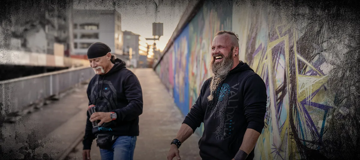 Zwei Männer in North Legendary Hoodies mit Wikinger-Print stehen vor einer Graffiti-Wand und lachen gemeinsam