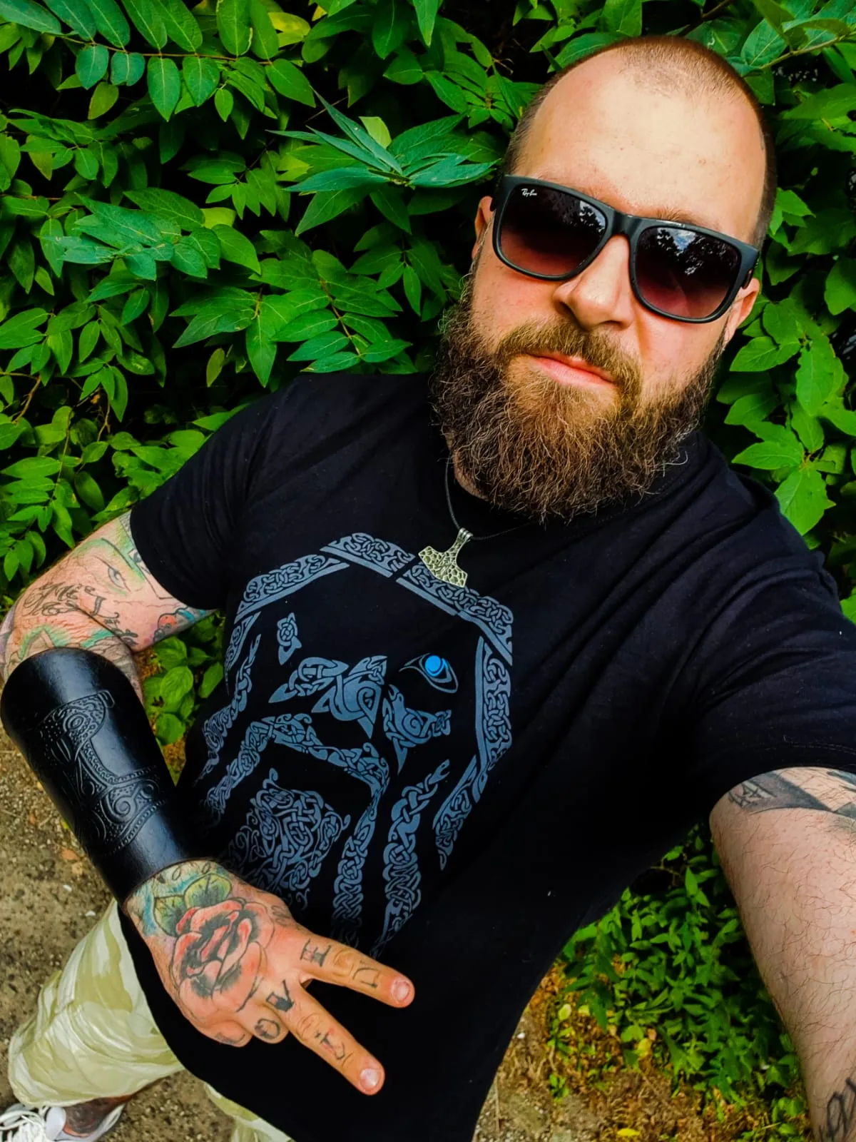 Tätowierter Mann mit Sonnenbrille und Bart vor grüner Hecke, trägt North Legendary Odin-Shirt mit blauem Auge