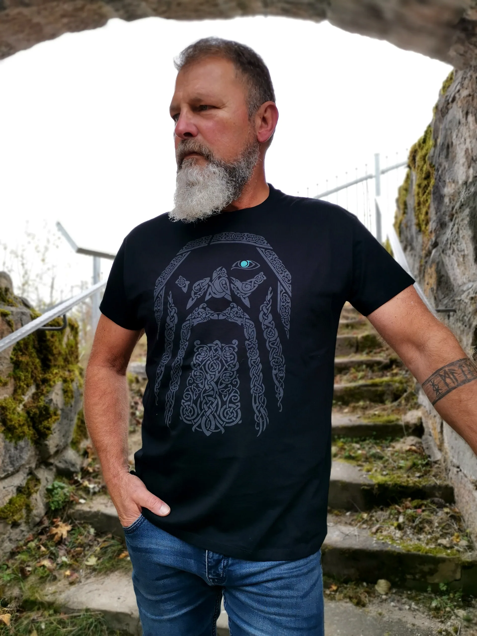 Mann mit grauem Bart und Odin-Shirt mit Wikinger-Kopf-Motiv im Bogen einer Steinmauer