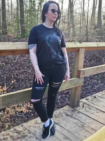 Frau mit Sonnenbrille trägt schwarzes North Legendary Hugin-und-Munin-Shirt auf einer Holzbrücke im Wald