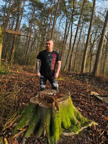 Mann im Berserker-Shirt mit rotem Print steht im Wald vor einem moosbewachsenen Baumstumpf