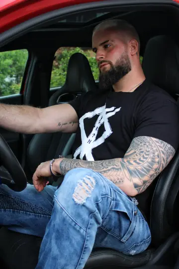 Tätowierter Mann mit Bart sitzt im Auto, trägt North Legendary Rune-Shirt mit weißem Print