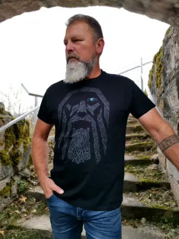 Mann mit grauem Bart und Odin-Shirt mit Wikinger-Kopf-Motiv im Bogen einer Steinmauer